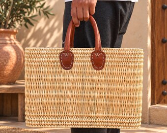 May include: A rectangular woven straw basket featuring a light tan natural fibre body with a vertical weave pattern. The basket includes two sturdy brown leather handles securely attached to the top edge with visible white stitching on leather tabs.