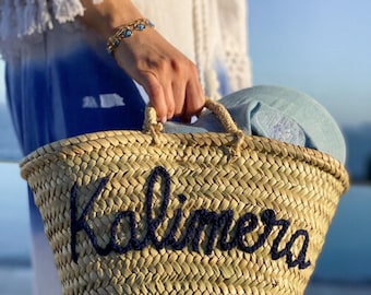 May include: A woven straw basket bag featuring two short handles and the word Kalimera embroidered in dark blue yarn across the front. The basket has a natural tan colour and a textured, braided construction with a wide, open top design.