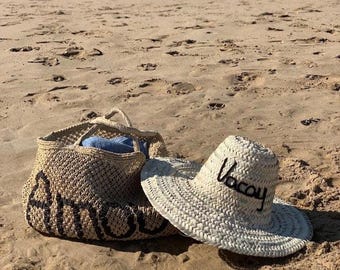 May include: A beige woven beach bag and sun hat on a sandy beach. The bag has black lettering that says "amour". The hat is white with the word "Vacay" embroidered in black.