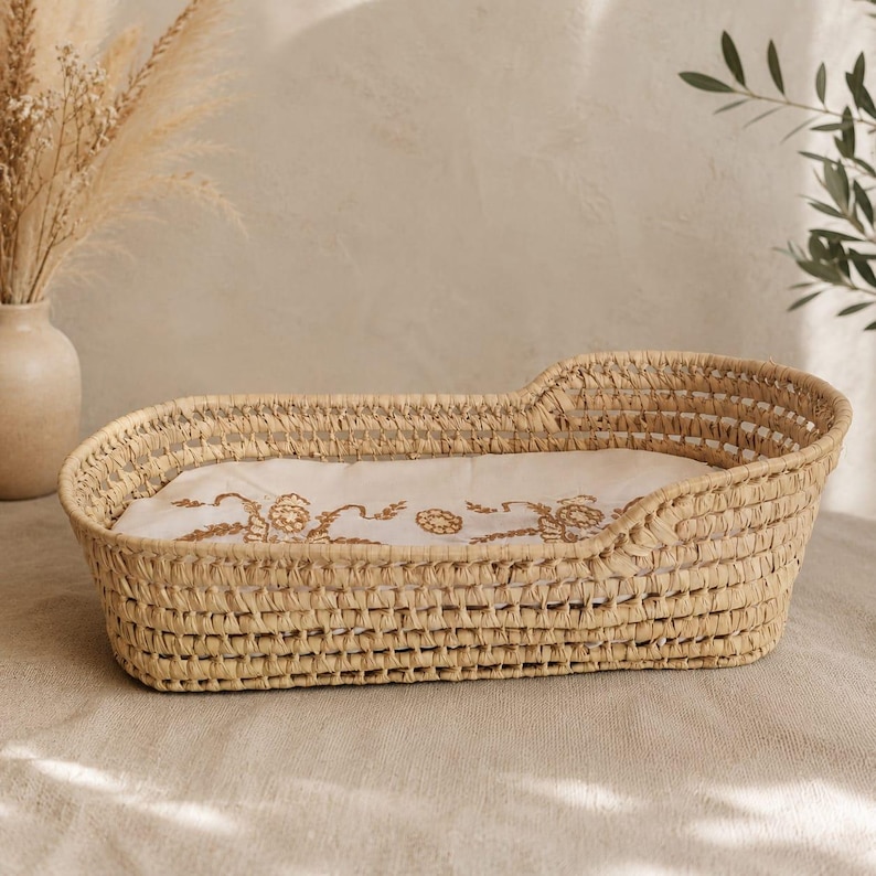 Puede incluir: Una cuna de mimbre tejido con un forro de tela bordada de color crema. La cuna tiene una forma curva con laterales elevados y una base rectangular. El forro de tela presenta un dise&ntilde;o floral en un tono c&aacute;lido y terroso.