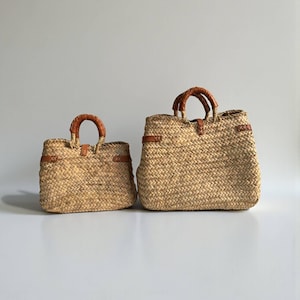 May include: Two woven straw tote bags of different sizes, each featuring a natural tan woven texture, two rounded brown leather handles, and brown leather strap accents with visible stitching details across the top edges of the bags.