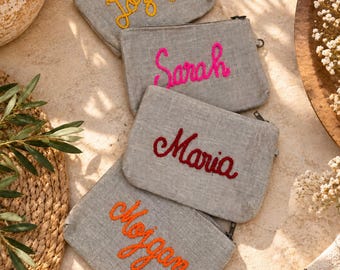 May include: Four rectangular grey linen pouches with top zippers. Each pouch features a name embroidered in a different colour thread: yellow for Jozefa, hot pink for Sarah, dark red for Maria, and bright orange for Mojgan in cursive script.