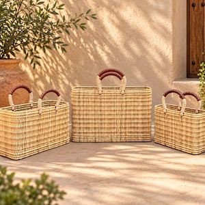 May include: Three rectangular woven baskets of varying sizes, each featuring a light tan natural fiber construction and two sturdy handles wrapped in dark brown leather. The baskets have a structured, flat-bottomed design with a consistent weave.