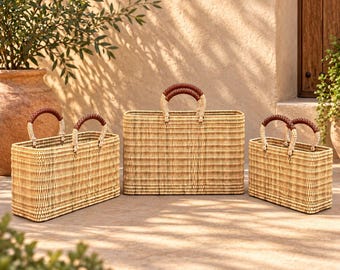 May include: Three rectangular woven baskets of varying sizes, each featuring a light tan natural fibre construction and two sturdy handles wrapped in dark brown leather. The baskets have a structured, flat-bottomed design with a consistent weave.