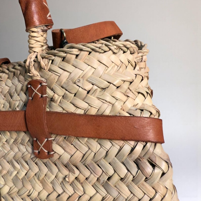 May include: A woven straw basket bag featuring tan leather straps and handles. The leather components are secured with white cross-stitch detailing. The bag has a natural light beige color and a textured, hand-woven pattern throughout the body.