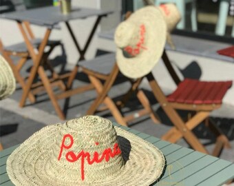 May include: A natural straw hat with a wide brim, embroidered with the word "Papins" in red. The hat sits on a green table, with other hats and outdoor seating in the background. A classic summer accessory, perfect for sunny days.