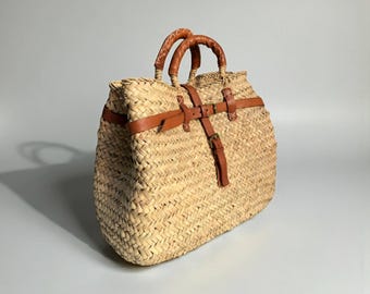 May include: Woven straw tote bag with brown leather handles and straps. The bag has a rectangular shape with a woven pattern and a buckle closure. The handles are curved and stitched, and the straps are secured with metal buckles.