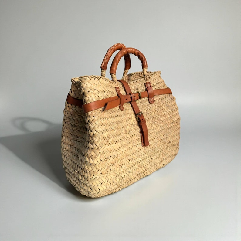 May include: A woven straw tote bag featuring a natural tan color with a textured herringbone pattern. The bag includes two braided leather handles and a brown leather strap closure system with brass-toned metal buckles across the front.