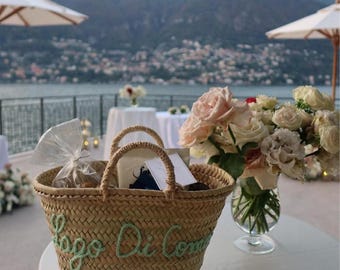 May include: A woven straw basket with two handles featuring the embroidered green text Lago Di Como 9/6/26 on the front. The basket contains a clear plastic bag and white paper items, positioned next to a glass vase filled with light pink roses.