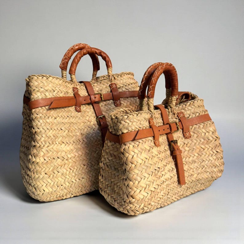 May include: Two woven straw tote bags featuring tan leather straps, brass buckles, and wrapped leather handles. The bags have a textured, braided straw construction with a horizontal leather belt detail across the front and a vertical strap.