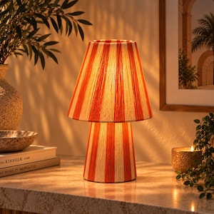 May include: A table lamp featuring a conical shade and a cylindrical base, both wrapped in a textured material with alternating vertical stripes in bright orange and cream. The lamp emits a warm, soft glow from within, illuminating its surface.