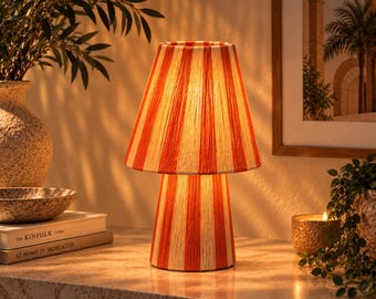May include: A table lamp featuring a conical shade and a cylindrical base, both wrapped in a textured material with alternating vertical stripes in bright orange and cream. The lamp emits a warm, soft glow from within, illuminating its surface.