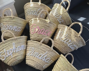 May include: A collection of woven straw baskets with handles, each featuring a name embroidered in white or pink yarn. The names visible on the baskets include Deanna, Heather, Grace, Olivia, Stephanie, Lindsay, and a partial name St.