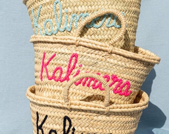 May include: Three woven straw tote bags stacked. Each bag features the word "Kalimera" embroidered in different colours: light blue, pink, and black. The bags have handles and are set against a light blue backdrop.