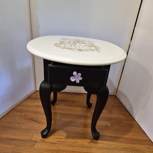 May include: An oval side table with a white top featuring a gold-toned decorative emblem. The base and single drawer are painted black, and the drawer is accented with a light purple flower-shaped knob. The table stands on four curved legs.