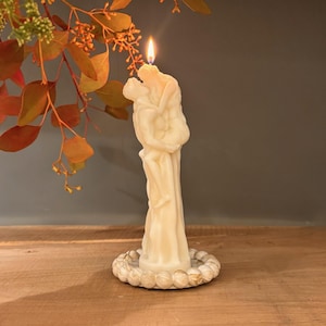Puede incluir: Una vela encendida de color marfil con forma de figuras abrazándose. La vela está sobre una base circular decorativa. La llama parpadea brillantemente. En el fondo, follaje otoñal con hojas naranjas y verdes. La vela mide unos 15 cm de altura.