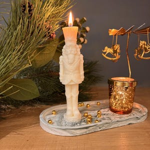 Op de afbeelding: Een brandende, gebroken witte notenkrakerkaars staat op een marmeren dienblad met gouden accenten. Een kleine, goudkleurige carrousel met schommelpaardfiguren en een goudkleurige votiefkaarsenhouder staan ook op de dienblad. Dennentakken en gouden kralen maken de feestelijke scène compleet.