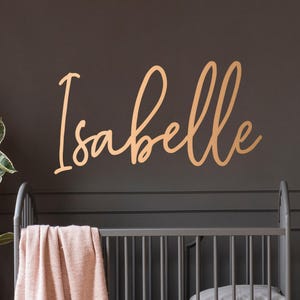 May include: A nursery scene with a wooden crib, a pink blanket, and a wooden sign that says "Isabelle". A teddy bear sits on a wooden dresser next to a potted plant. A pink lamp and a small table with a pink phone complete the decor.