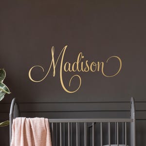 May include: A nursery scene with a gray crib, a pink blanket, and a wooden dresser. A teddy bear sits on the dresser next to a potted plant. The name "Madison" is written in gold script on the dark gray wall.