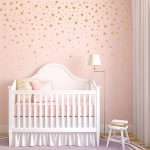 May include: A white crib with a pink mattress and bedding, set against a pink wall adorned with gold star decals. A floor lamp and small stool are also in the room, creating a soft, inviting nursery setting.