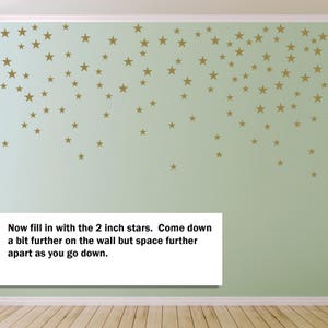 May include: A light green wall adorned with numerous gold-colored star decals. The stars vary in size and are arranged in a cascading pattern. Text on the image reads: "Now fill in with the 2 inch stars. Come down a bit further on the wall but space further apart as you go down."
