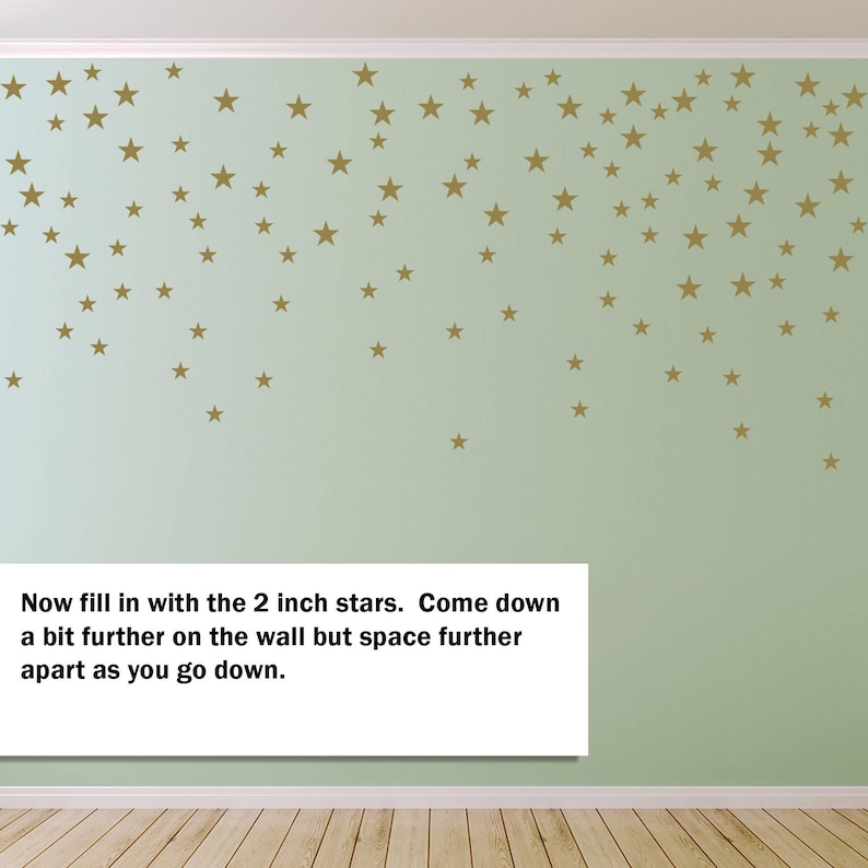May include: A light green wall adorned with numerous gold-colored star decals. The stars vary in size and are arranged in a cascading pattern. Text on the image reads: "Now fill in with the 2 inch stars. Come down a bit further on the wall but space further apart as you go down."