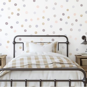 Puede incluir: Una habitaci&oacute;n con una cama de hierro forjado, ropa de cama blanca y una colcha a cuadros beige y blanca. La pared blanca est&aacute; adornada con lunares de acuarela en gris, beige y rosa. Tambi&eacute;n se ven una mesita de noche de madera y una l&aacute;mpara.