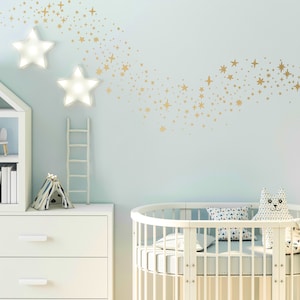 May include: A white cot with a white and blue patterned mattress and a white and blue patterned pillow. A white chest of drawers with three drawers is in the background. A white bookcase with a white cat figurine and a white bunny figurine is also in the background. The wall is light blue with gold star stickers in the shape of a shooting star.
