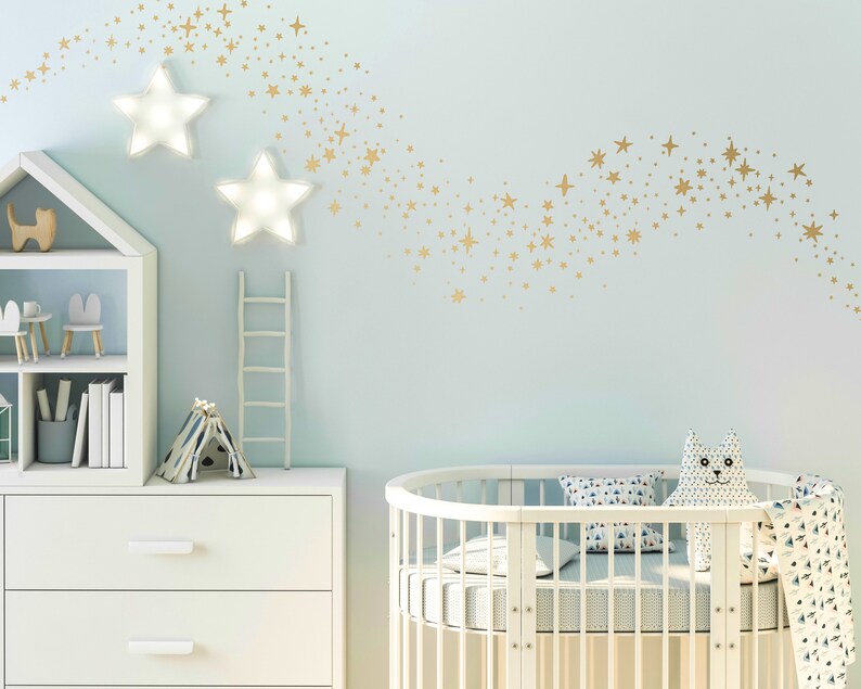 May include: A white cot with a white and blue patterned mattress and a white and blue patterned pillow. A white chest of drawers with three drawers is in the background. A white bookcase with a white cat figurine and a white bunny figurine is also in the background. The wall is light blue with gold star stickers in the shape of a shooting star.