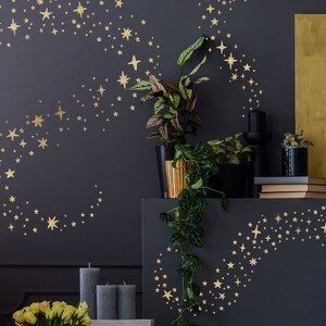May include: Gold star wall decals in a swirling pattern on a dark grey wall. The decals are arranged in a crescent shape and a swirling pattern. The wall is decorated with plants and candles.
