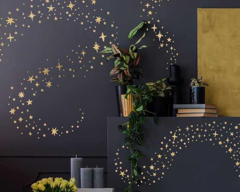 May include: Gold star wall decals in a swirling pattern on a dark grey wall. The decals are arranged in a crescent shape and a swirling pattern. The wall is decorated with plants and candles.