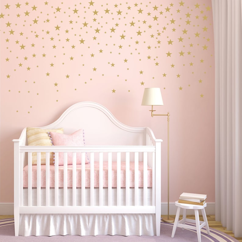 May include: A white crib with a pink mattress and bedding, set against a pink wall adorned with gold star decals. A floor lamp and small stool are also in the room, creating a soft, inviting nursery setting.