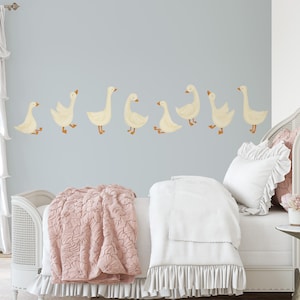 May include: A white bed with a pink quilted blanket and white ruffled bedding. The wall behind the bed has a row of white geese decals.