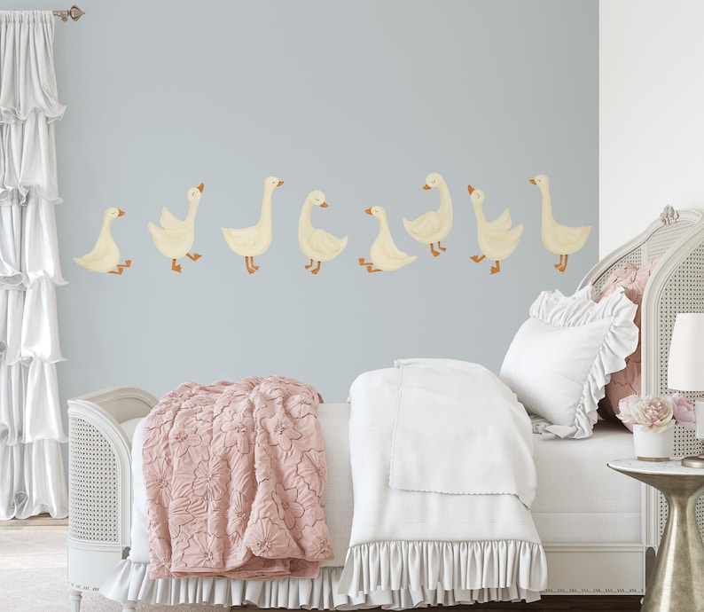 May include: A white bed with a pink quilted blanket and white ruffled bedding. The wall behind the bed has a row of white geese decals.