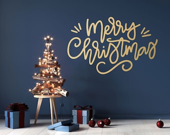 Puede incluir: Un árbol de Navidad rústico hecho de ramas y decorado con luces de cadena se encuentra sobre una mesa de madera frente a una pared azul. La pared tiene una inscripción dorada que dice "Feliz Navidad". Hay regalos envueltos y adornos rojos en el suelo.