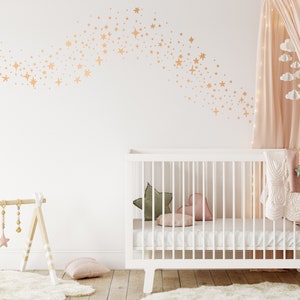May include: A white cot with a white mattress and a white blanket. There is a pink heart-shaped pillow and a green pillow in the cot. The cot is in a nursery with a white wall and a pink canopy. There are gold stars on the wall.