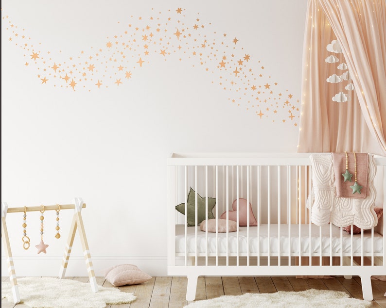 May include: A white cot with a white mattress and a white blanket. There is a pink heart-shaped pillow and a green pillow in the cot. The cot is in a nursery with a white wall and a pink canopy. There are gold stars on the wall.