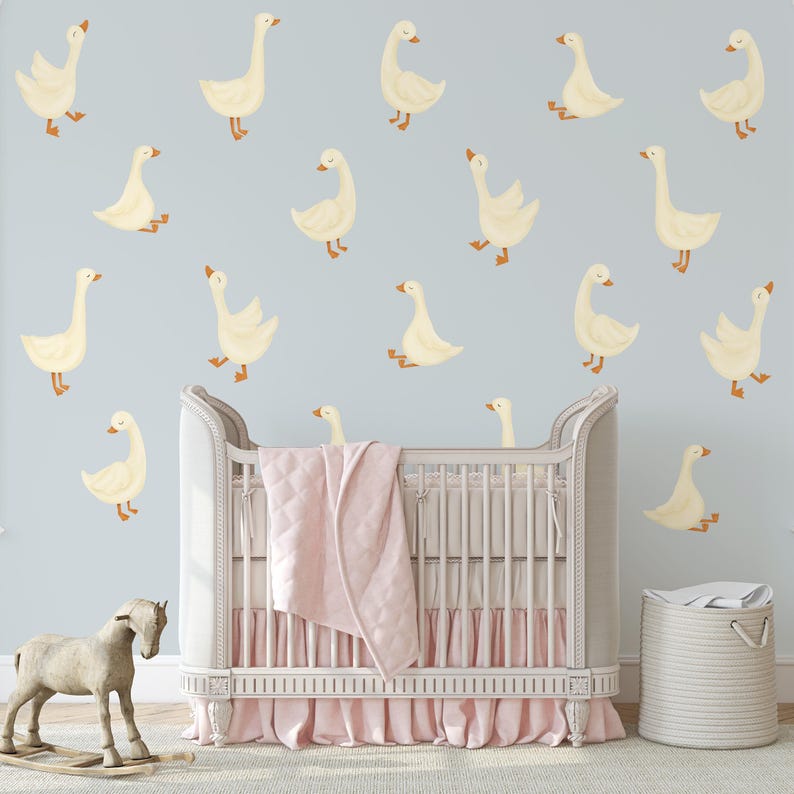 May include: A white crib with a pink ruffled skirt and a white blanket. The crib is decorated with yellow goose decals on a light blue wall. There is a rocking horse and a wicker basket in the room.