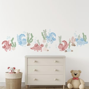 May include: A wall decal featuring a repeating pattern of cartoon axolotls in shades of pink and blue, with green seaweed and bubbles. The axolotls are smiling and swimming. A white dresser, a woven basket with stuffed animals, and a teddy bear are in the foreground.