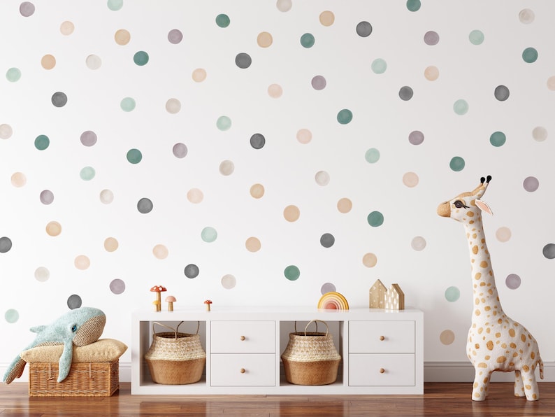 Puede incluir: Una pared blanca decorada con lunares de acuarela en varios colores, incluyendo gris, verde y beige. Un mueble blanco con cestas tejidas y un peluche de jirafa est&aacute;n en primer plano. Un peluche de ballena descansa sobre una cesta de mimbre.