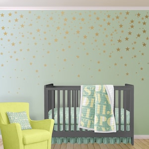 May include: A nursery scene with a gray crib, a lime green armchair, and a wall decorated with gold star decals. The crib has a matching crib skirt and a quilt with large letter designs. A decorative pillow rests on the armchair.
