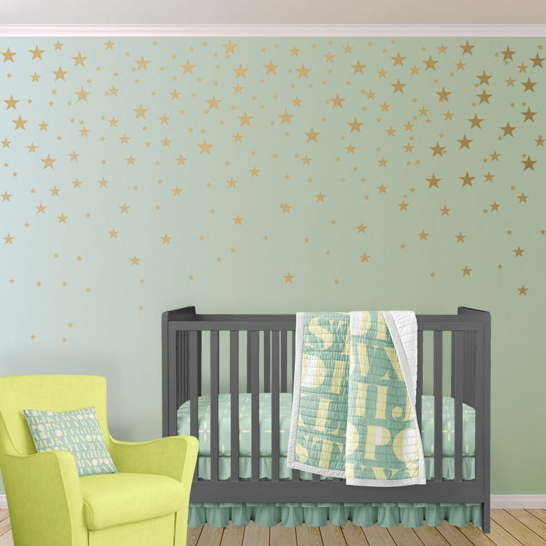 May include: A nursery scene with a gray crib, a lime green armchair, and a wall decorated with gold star decals. The crib has a matching crib skirt and a quilt with large letter designs. A decorative pillow rests on the armchair.