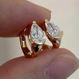 May include: A pair of gold hoop earrings, each featuring a pear-shaped, clear gemstone. The earrings are held up, showcasing their design and the way they would hang. The gold setting complements the sparkle of the stones.