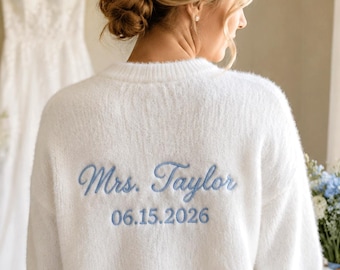 Könnte beinhalten: Ein weißer, flauschiger Langarmpullover mit hellblauer Stickerei auf der Rückseite, die Mrs. Taylor und das Datum 06.15.2026 zeigt. Der Pullover hat einen Rundhalsausschnitt und eine weiche, strukturierte Strickoberfläche, Rückansicht.