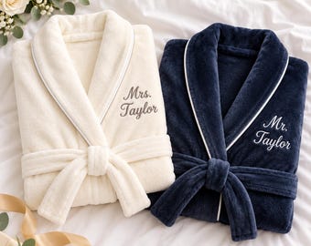 May include: A cream-coloured plush robe with white piping and the embroidered text Mrs. Taylor, alongside a navy blue plush robe with white piping and the embroidered text Mr. Taylor. Both robes feature a shawl collar and a matching tie belt.