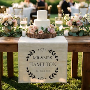 May include: A wedding table setting features a two-tiered white cake on a wooden base, with floral arrangements and candles. A linen table runner displays the text "MR.&MRS. HAMILTON" and the date 06.08.2026.