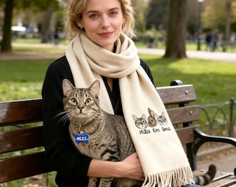 May include: A cream-colored scarf with a fringed edge, featuring embroidered cat faces and the names "Miles Kim Sena." A tabby cat with a blue "Miles" tag sits on a person's lap. The scene is set outdoors.