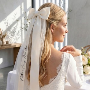 May include: Ivory bridal veil with a large bow and long ribbons. The veil is attached to the hair with a large bow. The veil has the text "Future Mrs. Marta" printed on it. The bride is wearing a white dress with lace detailing.