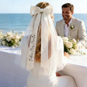 Pode incluir: Uma noiva usando um véu branco com um laço grande e o texto "Future Meg. Martin". O véu é combinado com um vestido branco de manga comprida. Um homem de terno bege sorri ao fundo. Há flores na mesa.
