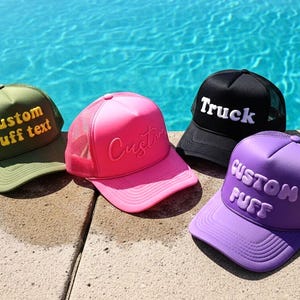 Puede incluir: Cuatro gorras de camionero en verde oliva, rosa, negro y morado. Cada gorra tiene una parte trasera de malla y una visera curva. La gorra verde oliva tiene "Custom Puff text" en amarillo. La gorra rosa tiene "Custom" en rosa. La gorra negra tiene "Truck" en blanco.